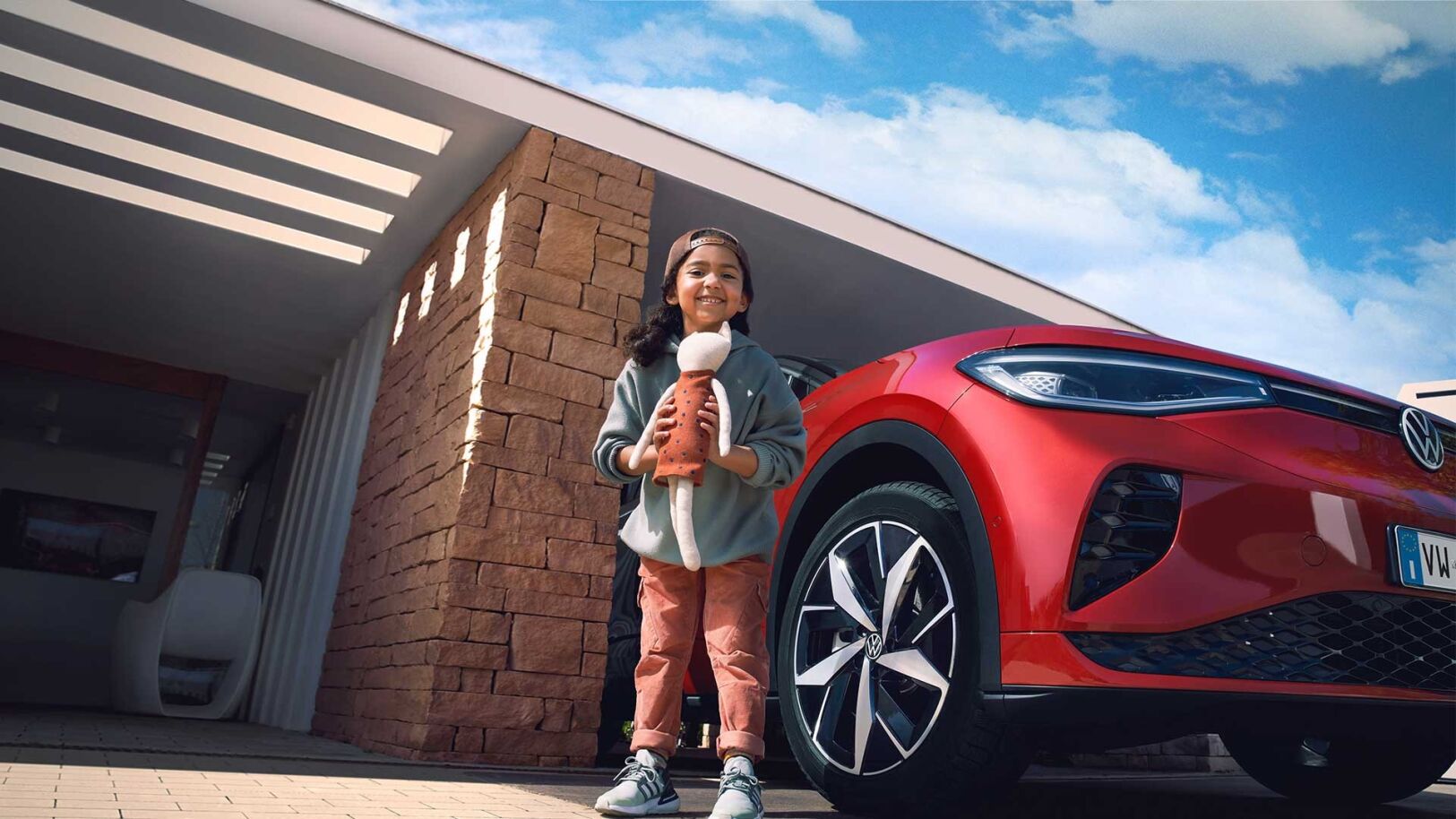 Inquadratura ravvicinata dal basso di una bambina sorridente mentre tiene un mano un pupazzo e dietro di lei parcheggiata in un garage una vettura Volkswagen.